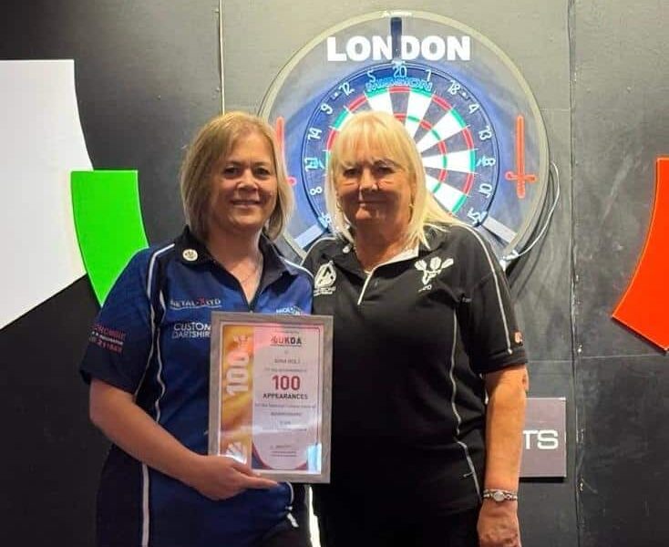 Nina receives award for 100 darts appearances, holding framed certificate.