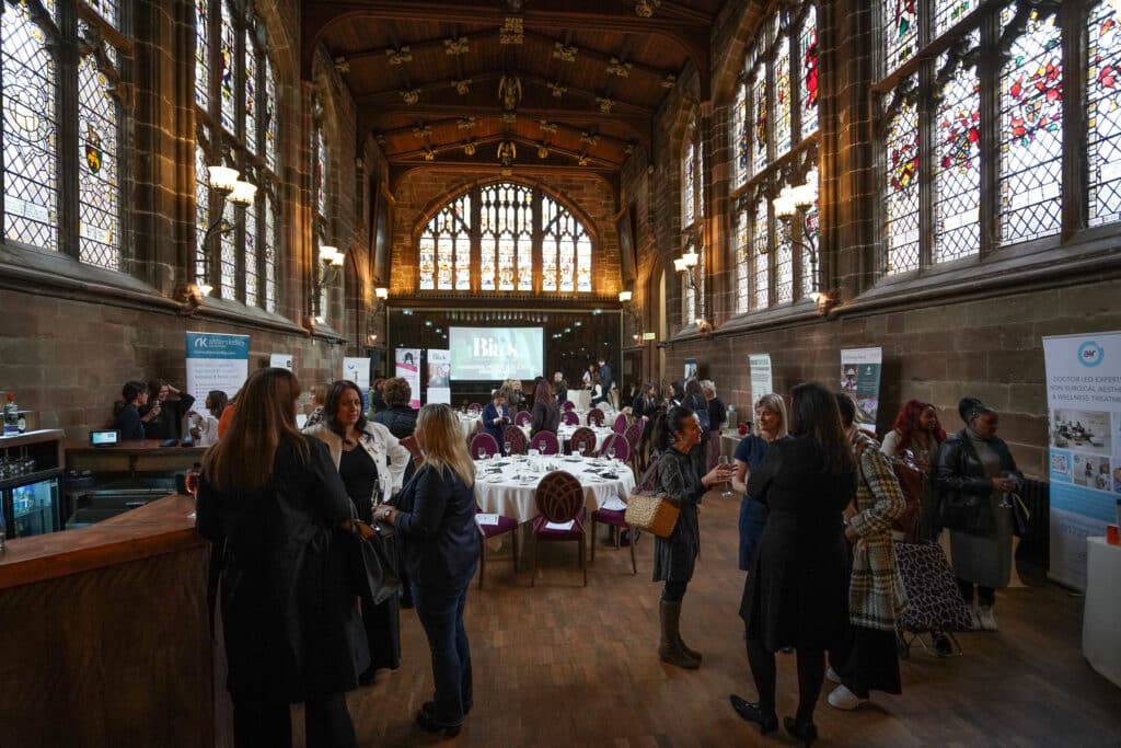 Networking event attendees converse in a hall with arched windows and a projector screen