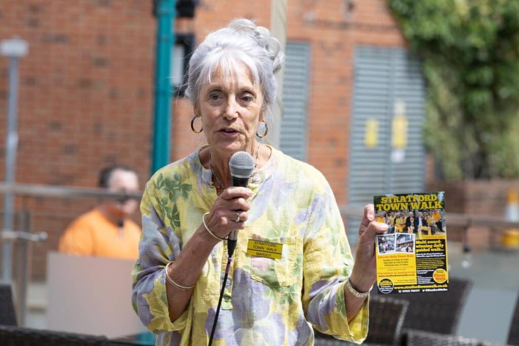 Woman with microphone and flyer for Stratford Town Walk, with text "Stratford Town Walk