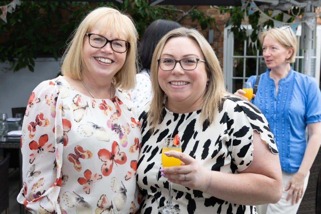 Two smiling women in casual attire holding drinks at an outdoor gathering