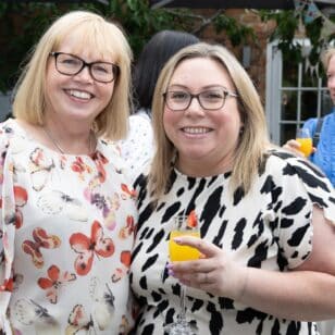 Two smiling women in casual attire holding drinks at an outdoor gathering