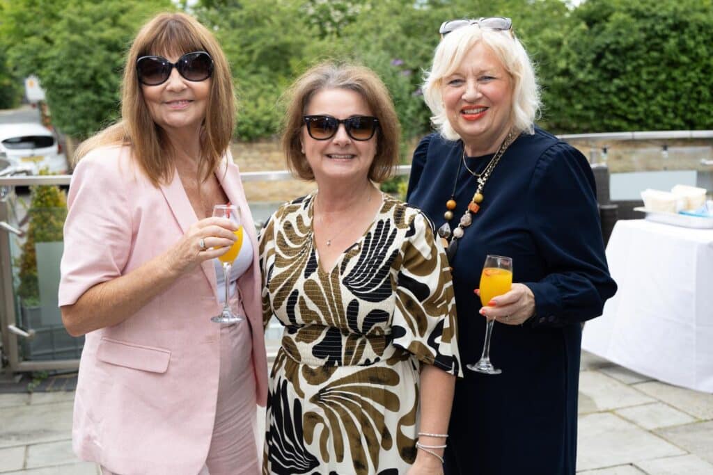 Three women in elegant attire holding drinks at an outdoor event.