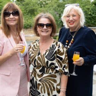 Three women in elegant attire holding drinks at an outdoor event.